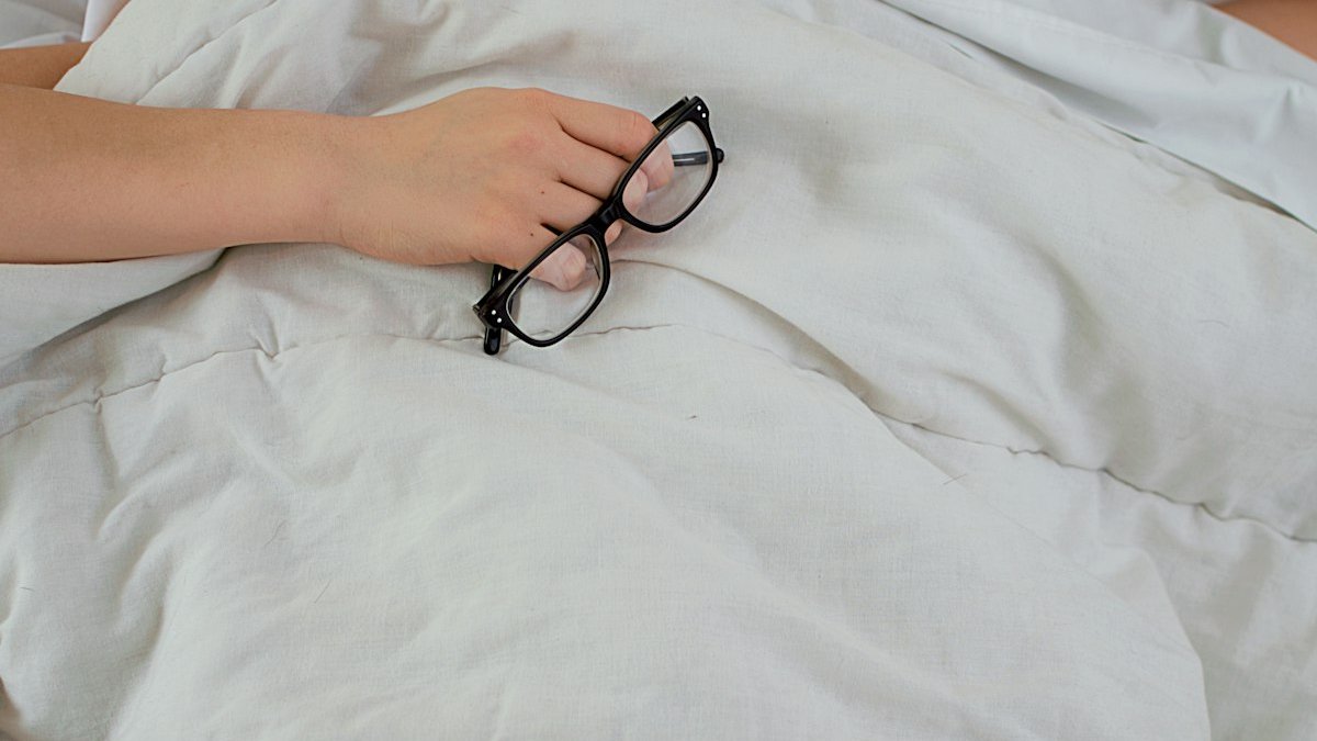 Person lying on bed while covering face with pillow and holding eyeglasses. — a Sleep Finally guide to Things to Do Before Bed Instead of Scrolling.