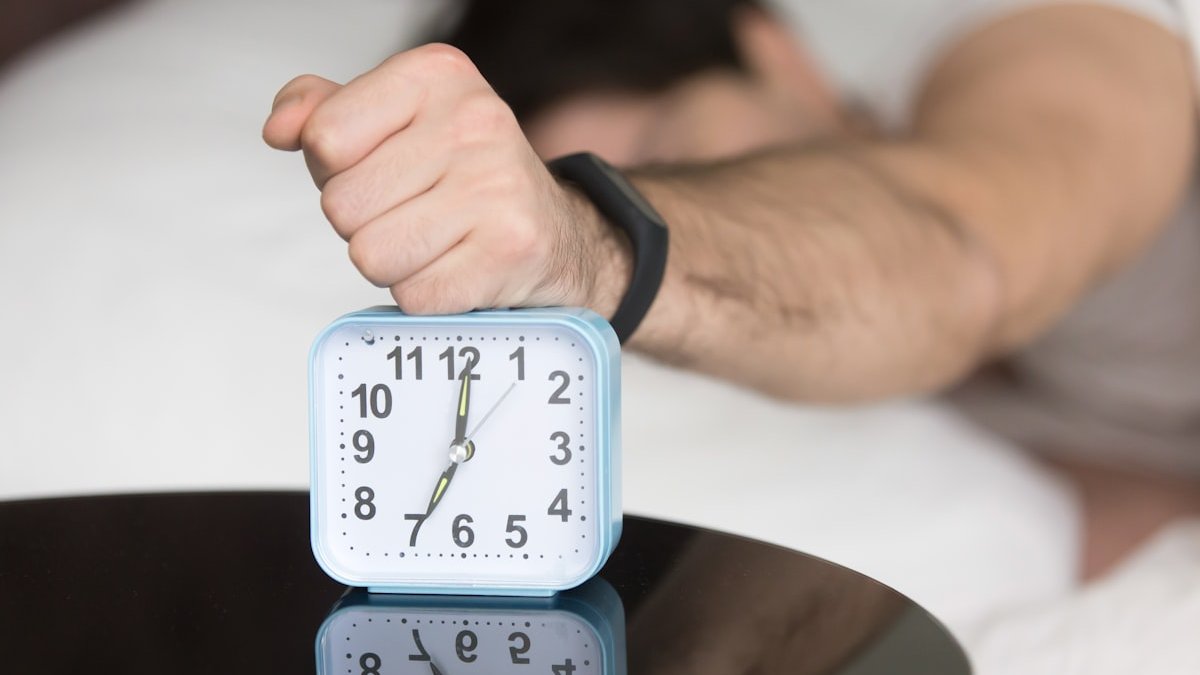 A man laying in bed with a clock on top of him. — a Sleep Finally guide to Waking at the Same Time Every Night: Causes Explained.
