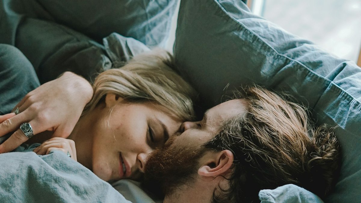 Man kissing woman's forehead while lying on bed. — a Sleep Finally guide to When Partners Keep Different Sleep Schedules.
